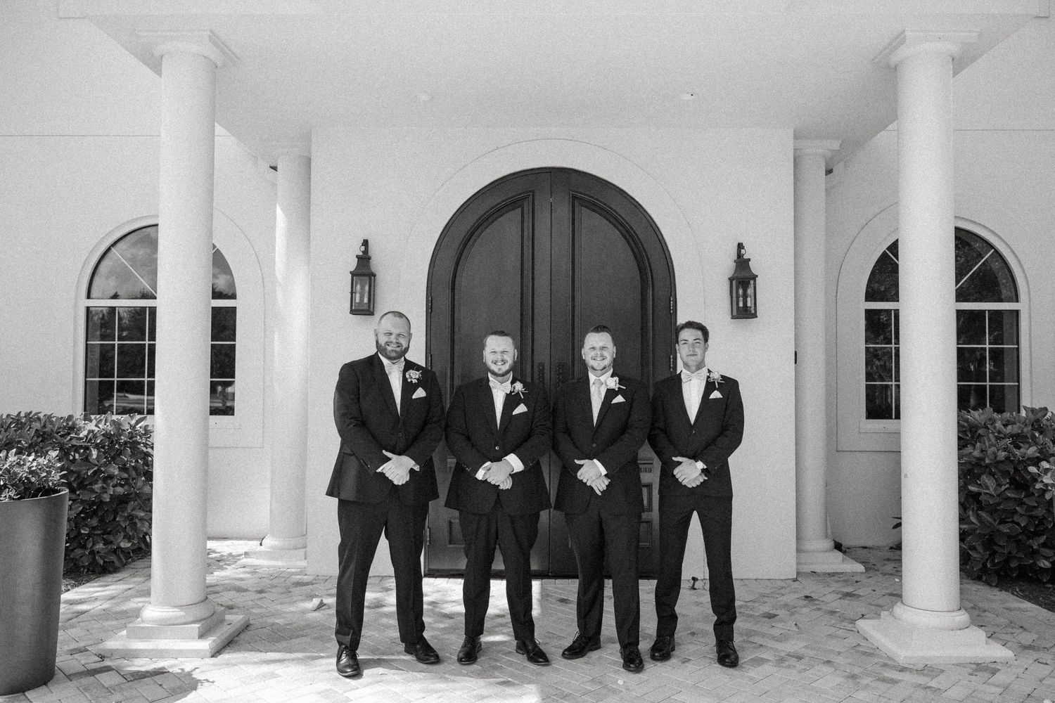 Five groomsmen in black suits stand in a row against a white columned exterior of a building.