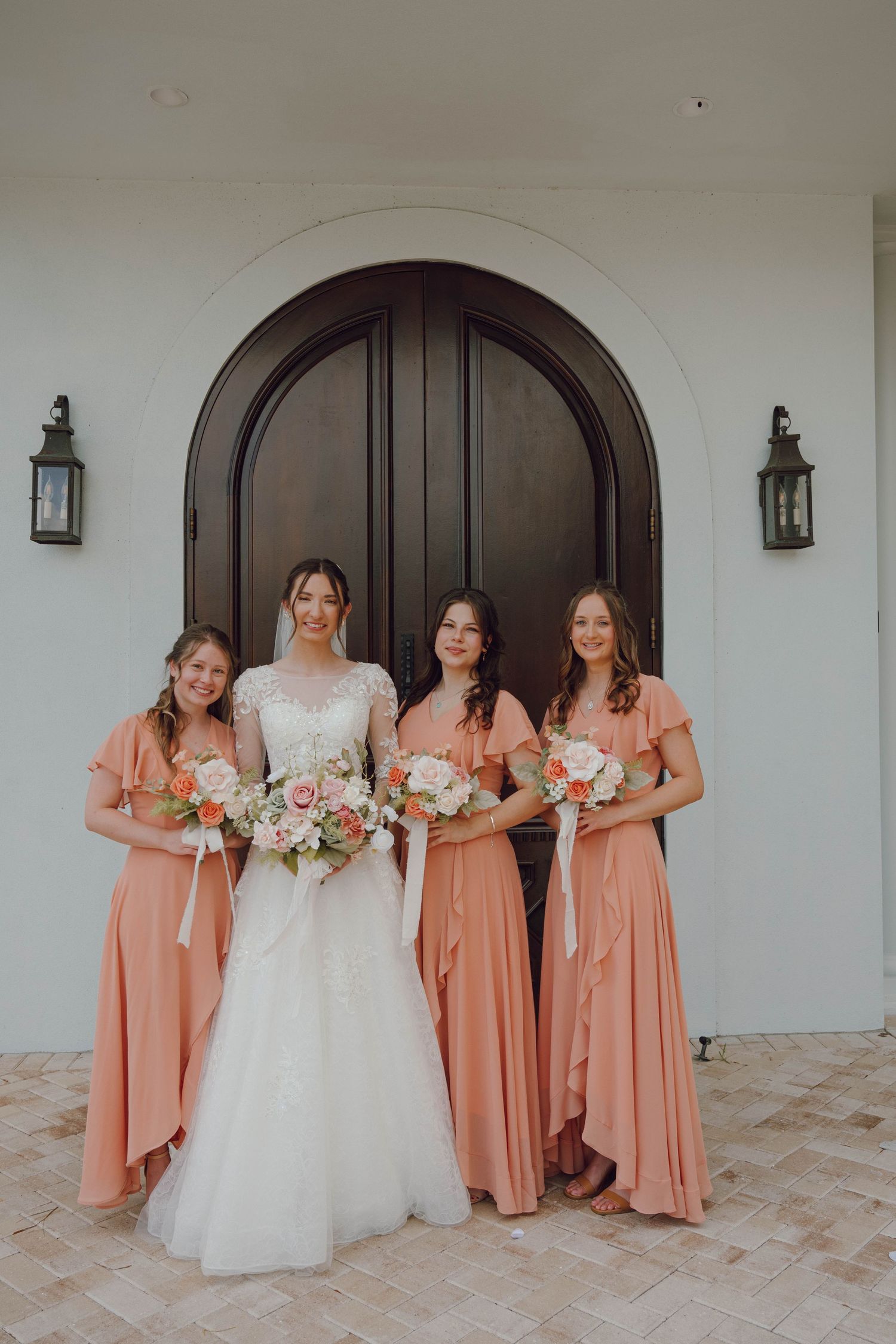 A bridal party poses in coral dresses alongside a bride in white against an arched doorway.