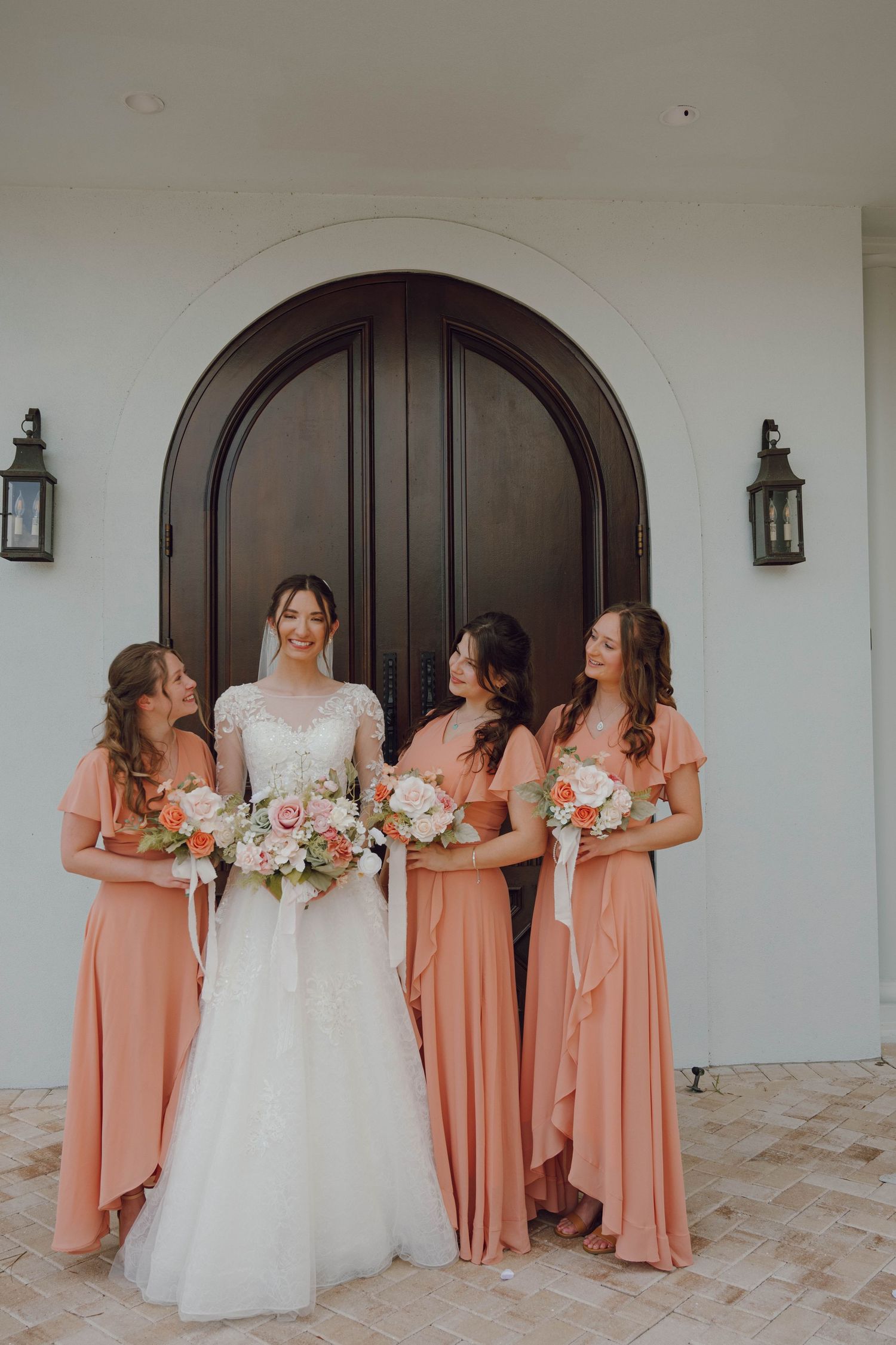 Bridesmaids in peach dresses gather around bride in white gown near arched doorway.