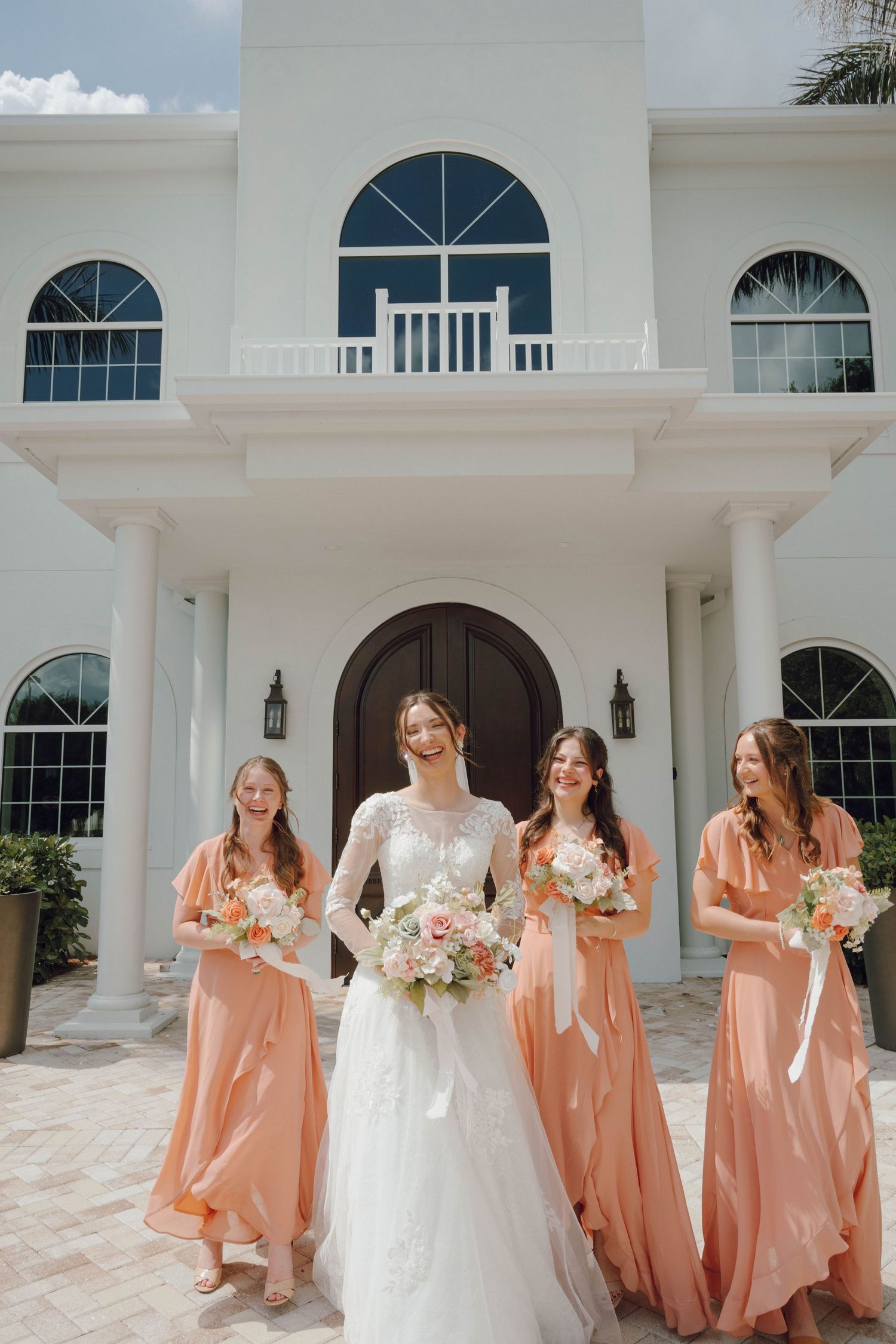 Wedding party poses outside white mansion with bridesmaids in coral dresses and bride in white gown.