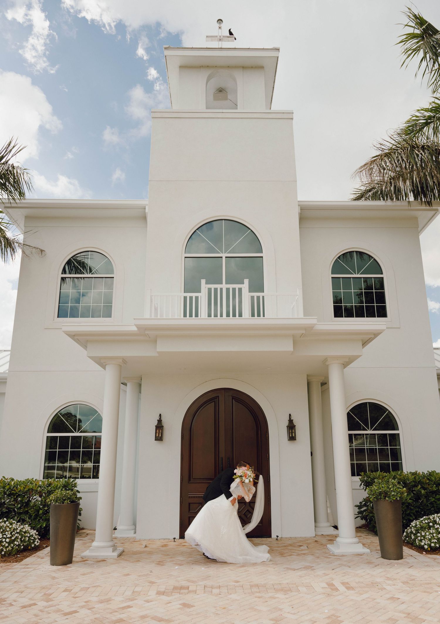 Elegant white church-style wedding venue with arched windows and a tall steeple tower against a blue sky with palm trees.