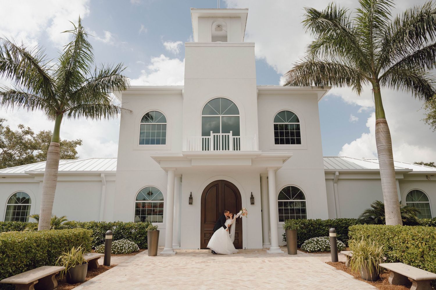 A luxurious white Mediterranean-style villa with palm trees and a grand entrance in a tropical setting.