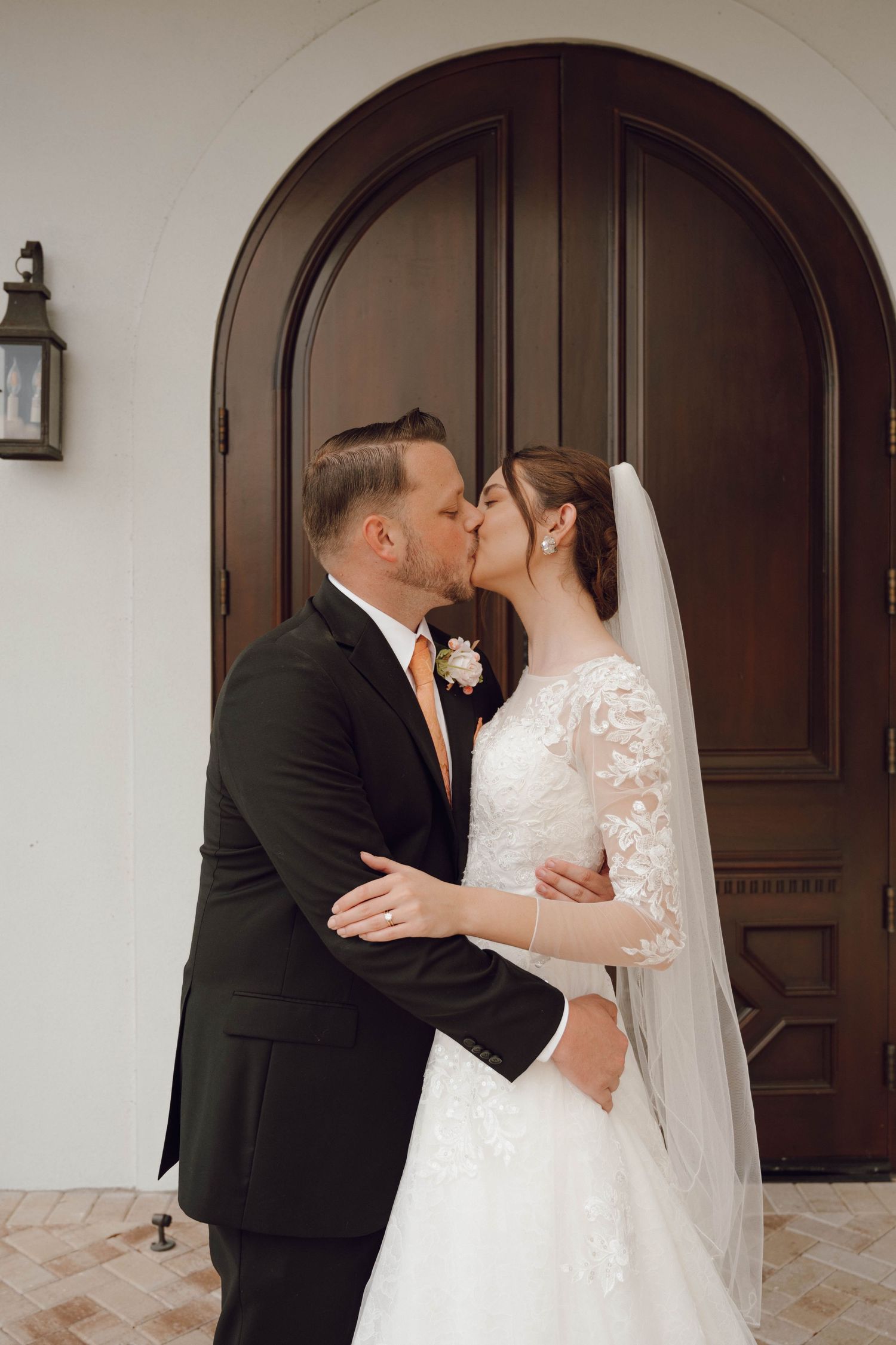 Romantic wedding moment captured in front of a grand wooden arched door at an elegant white chapel entrance.