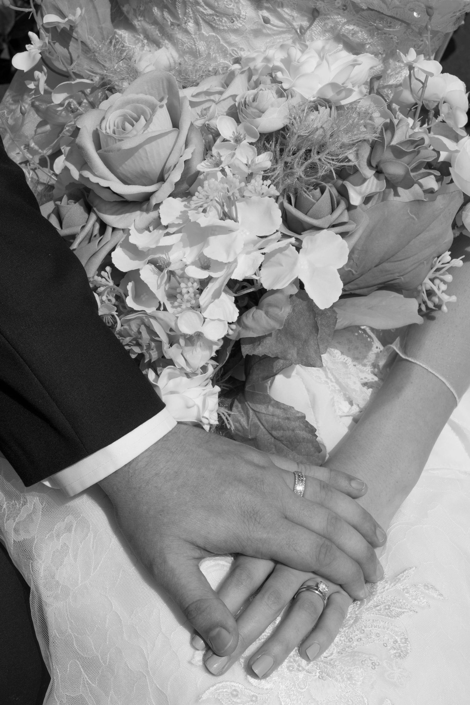 A close-up black and white photo of hands with wedding rings and a bouquet of roses and mixed flowers.