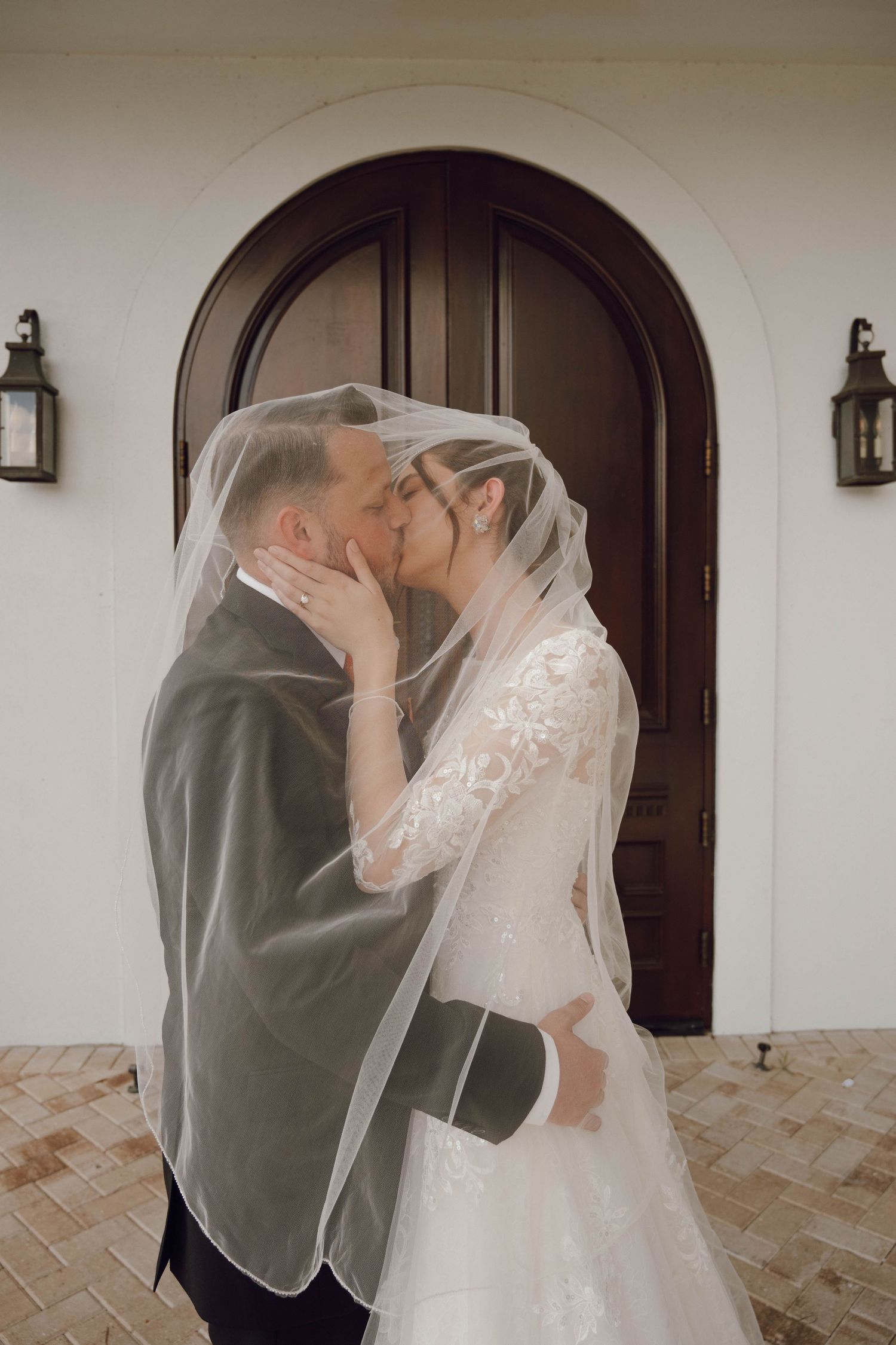 A romantic wedding kiss in front of an arched wooden door with lanterns on either side.