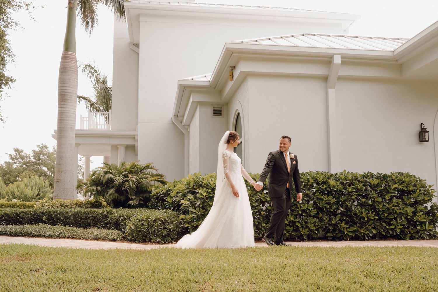 A couple in wedding attire stands together on a lawn outside a white Mediterranean-style building with palm trees.