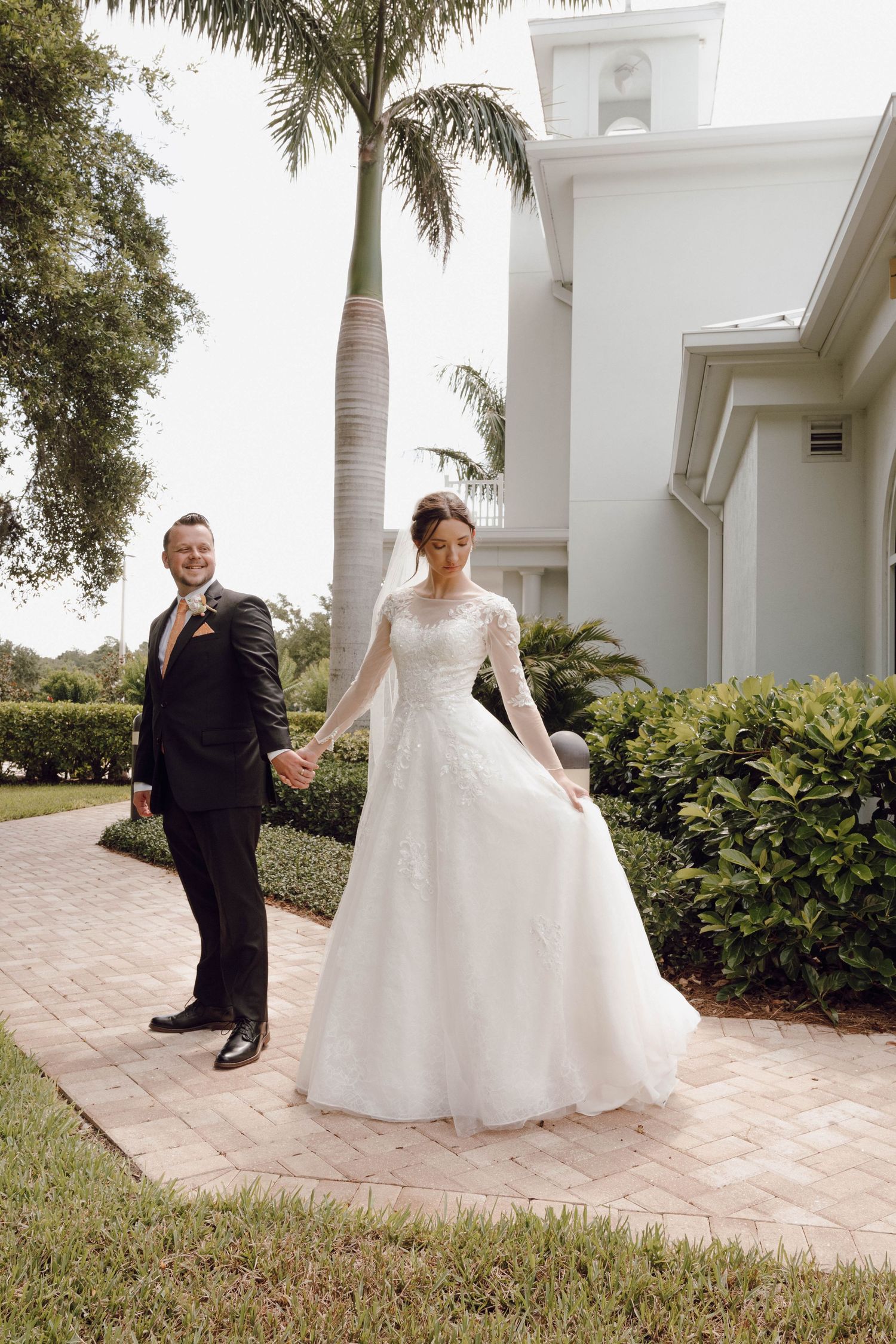 A couple in wedding attire poses on a pathway with palm trees and a white church building.