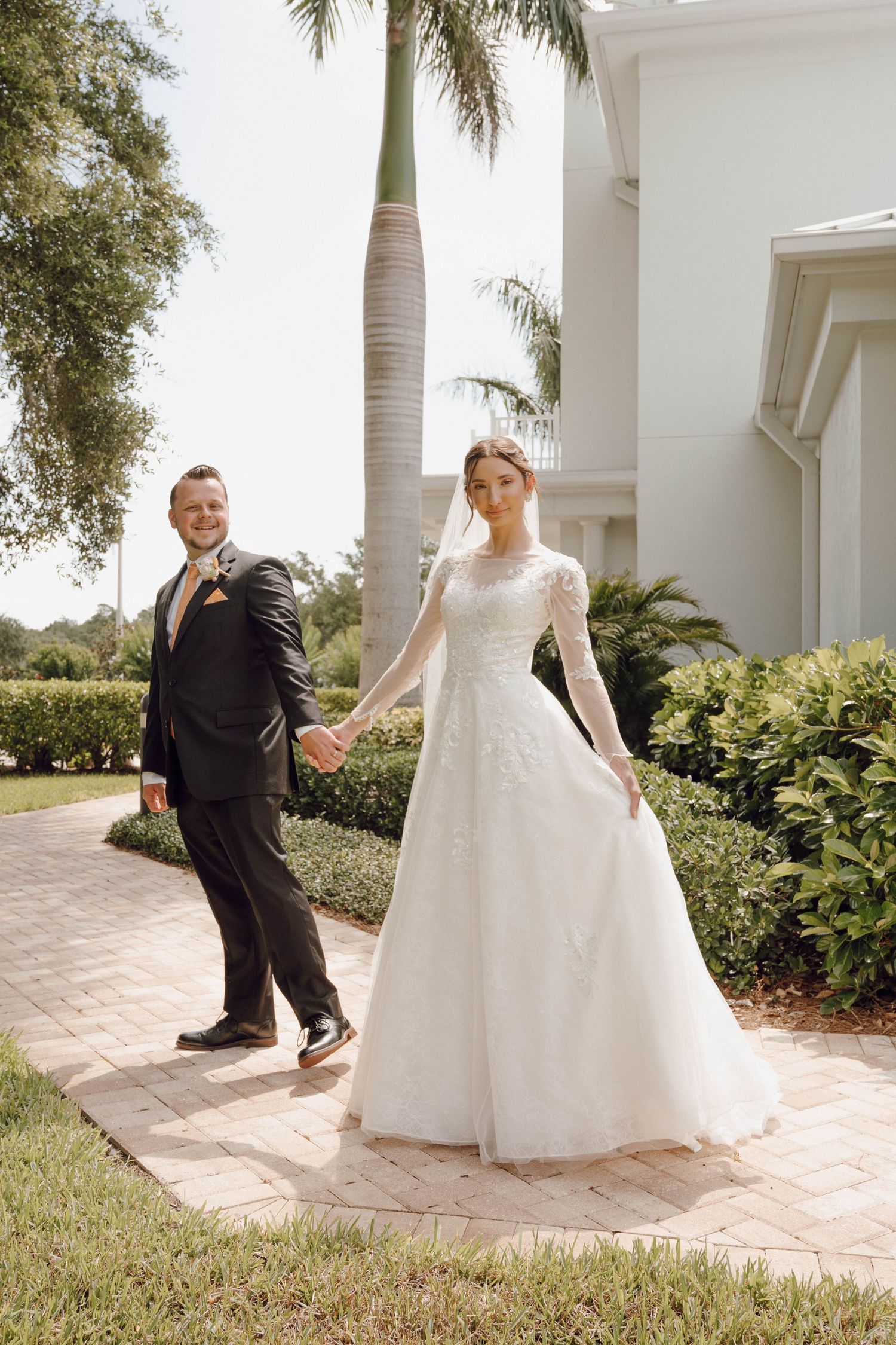A couple in wedding attire poses on a garden path with palm trees and white architecture.