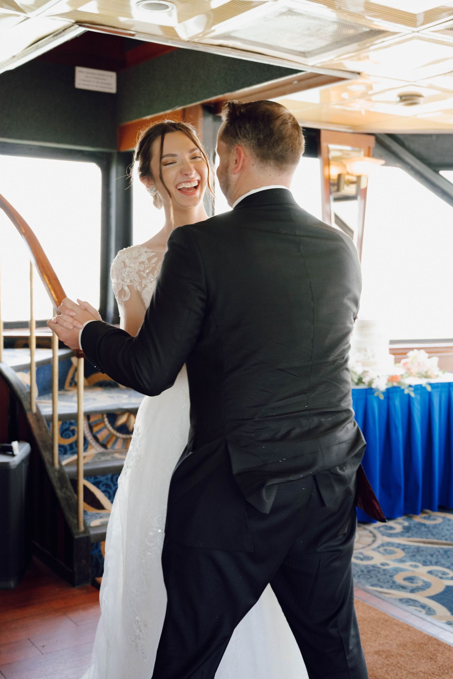 A couple in wedding attire share a joyful dance moment aboard a boat with blue seating visible in the background.