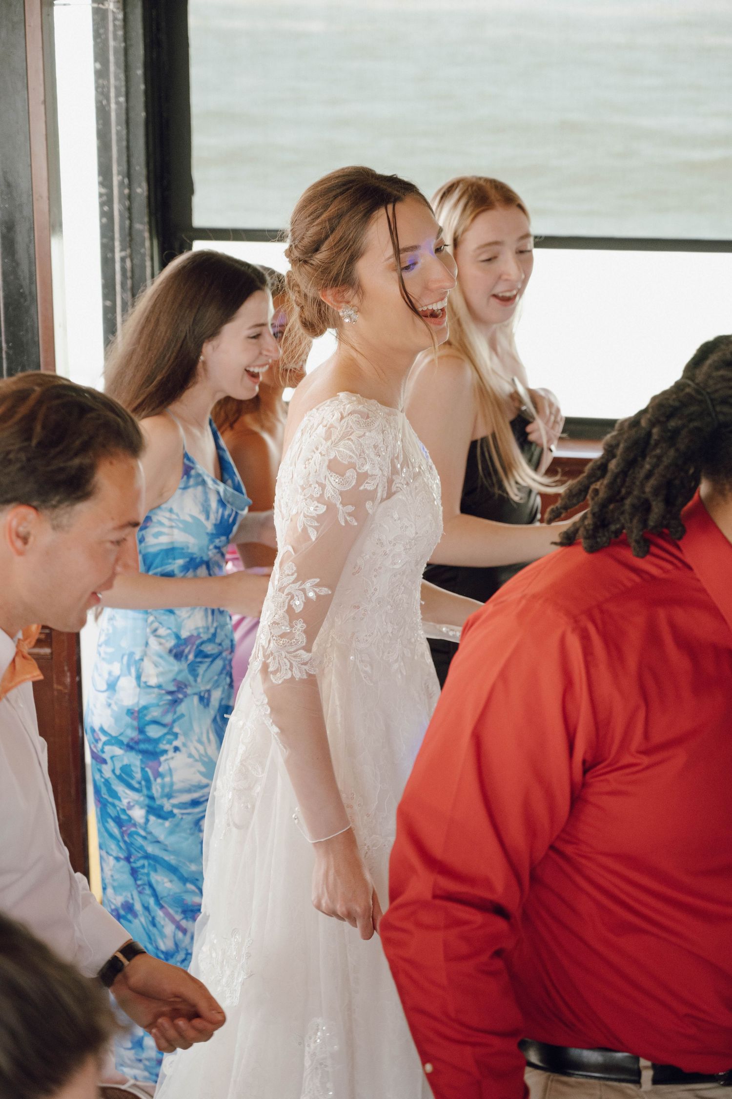 A joyful wedding party laughs together during a candid moment on what appears to be a bus or trolley.