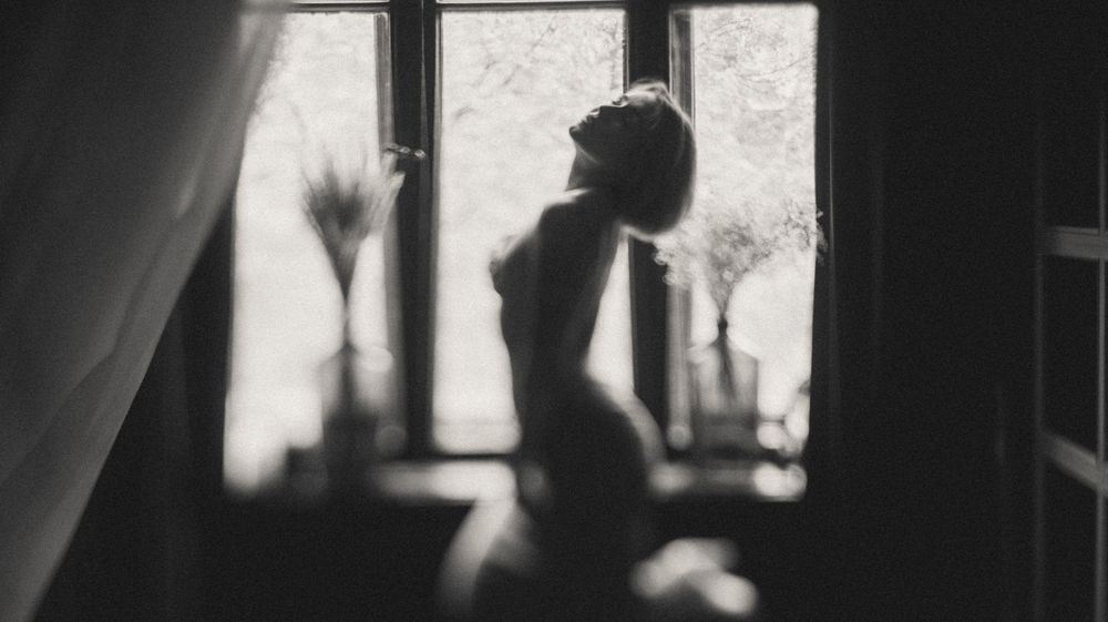 Artistic black and white silhouette of a person standing in a window frame with dramatic lighting and shadows.