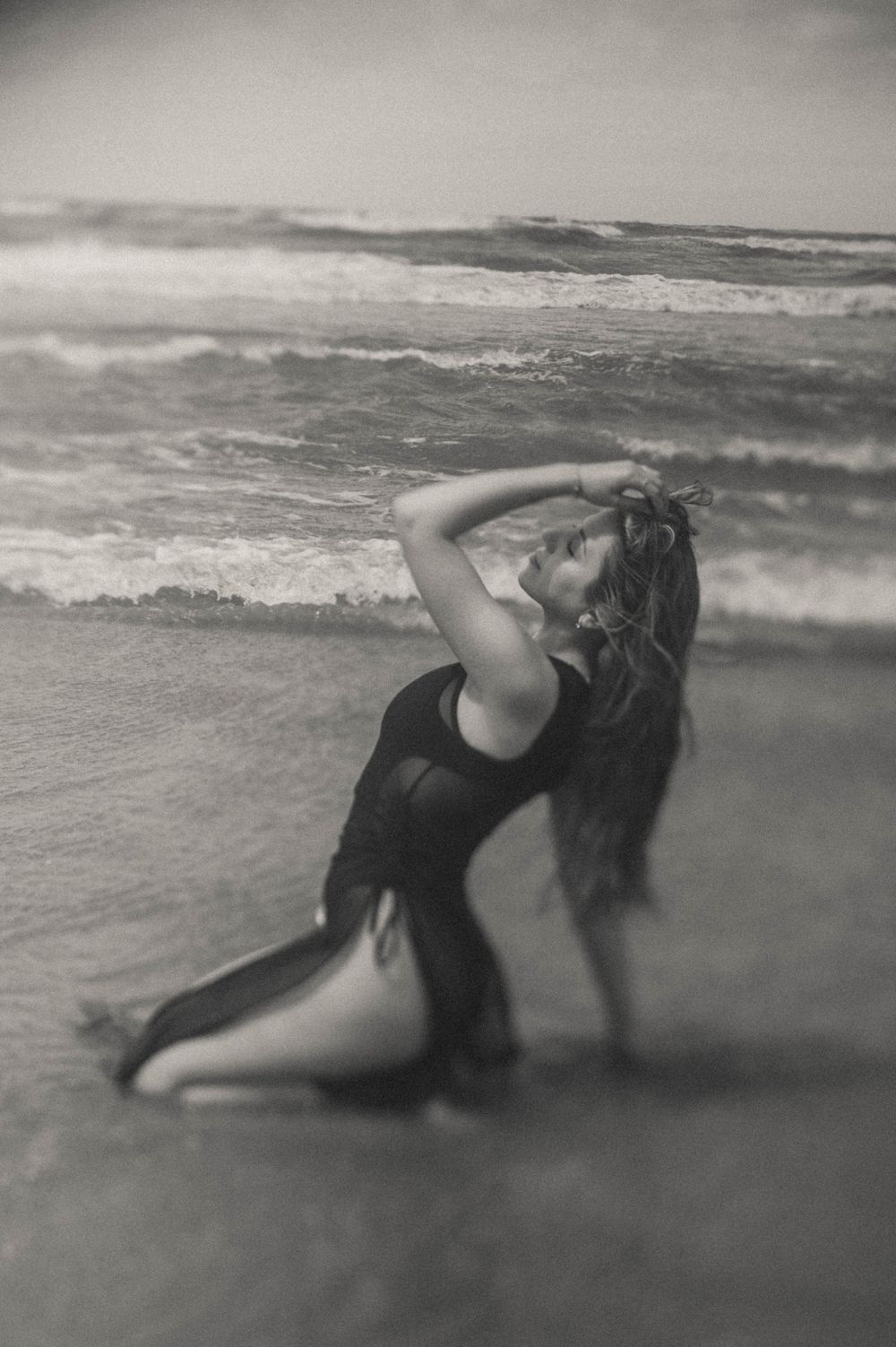 Artistic black and white boudoir photo of a woman moving gracefully in ocean waves at the shoreline.