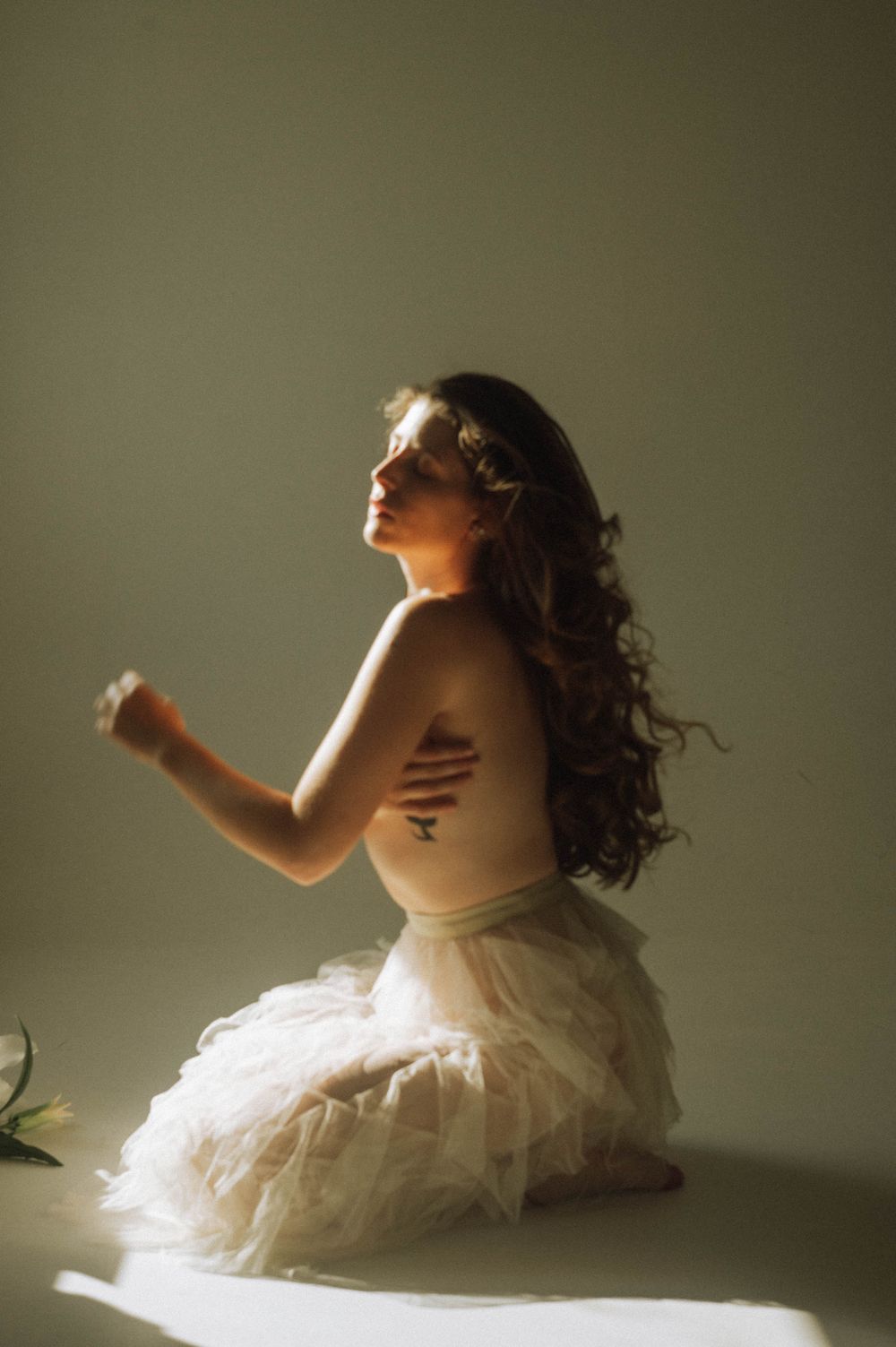 Artistic portrait of figure in white tulle skirt illuminated by dramatic side lighting in moody setting.
