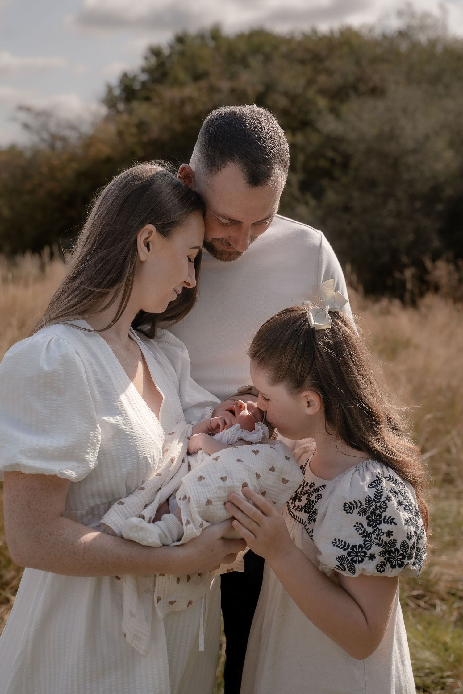 Family in white clothing sharing tender moment with newborn baby in grassy field during golden hour.