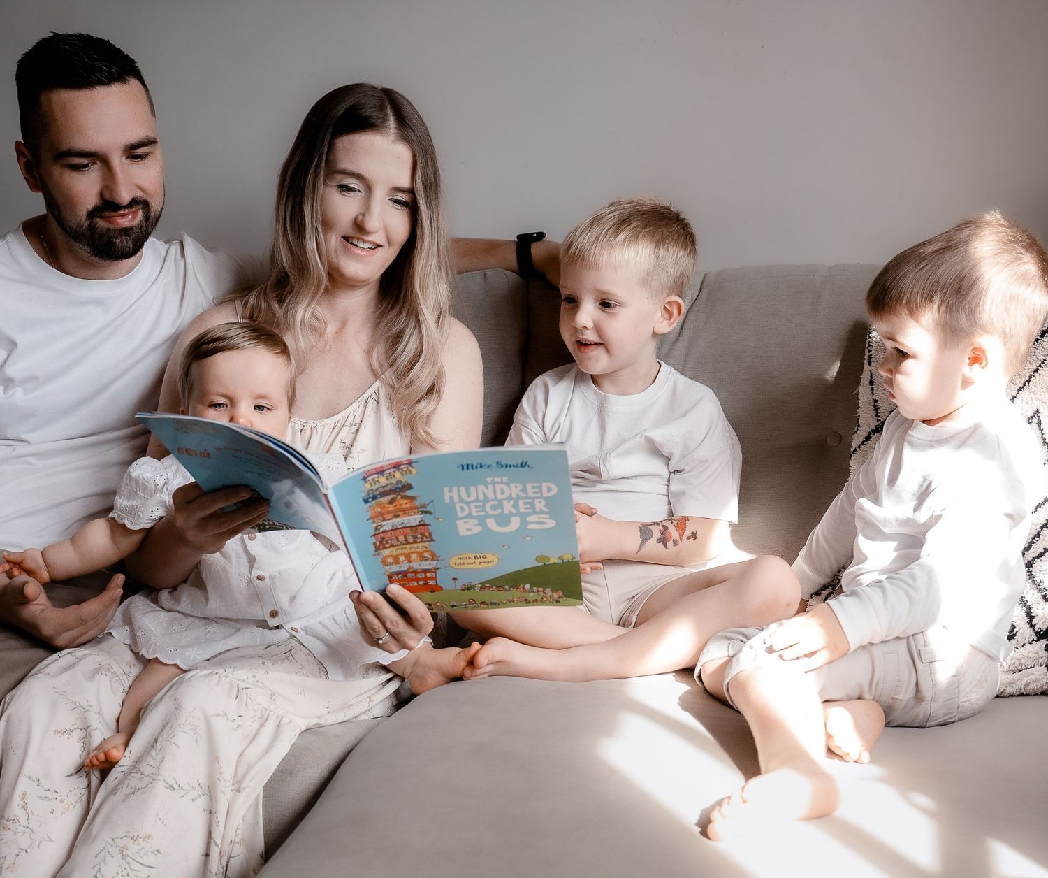 A family in white clothing sits together on a bed reading a book in warm natural light.