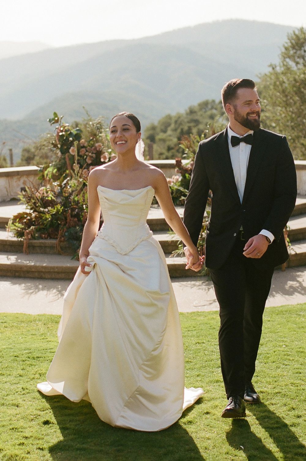 A couple walks on grass with mountains in the background during their wedding.