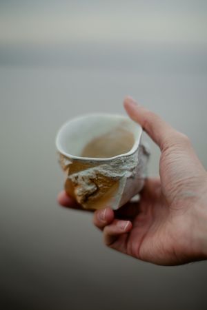 A hand holds a delicate white ceramic teacup with textured edges against a blurred gray background.