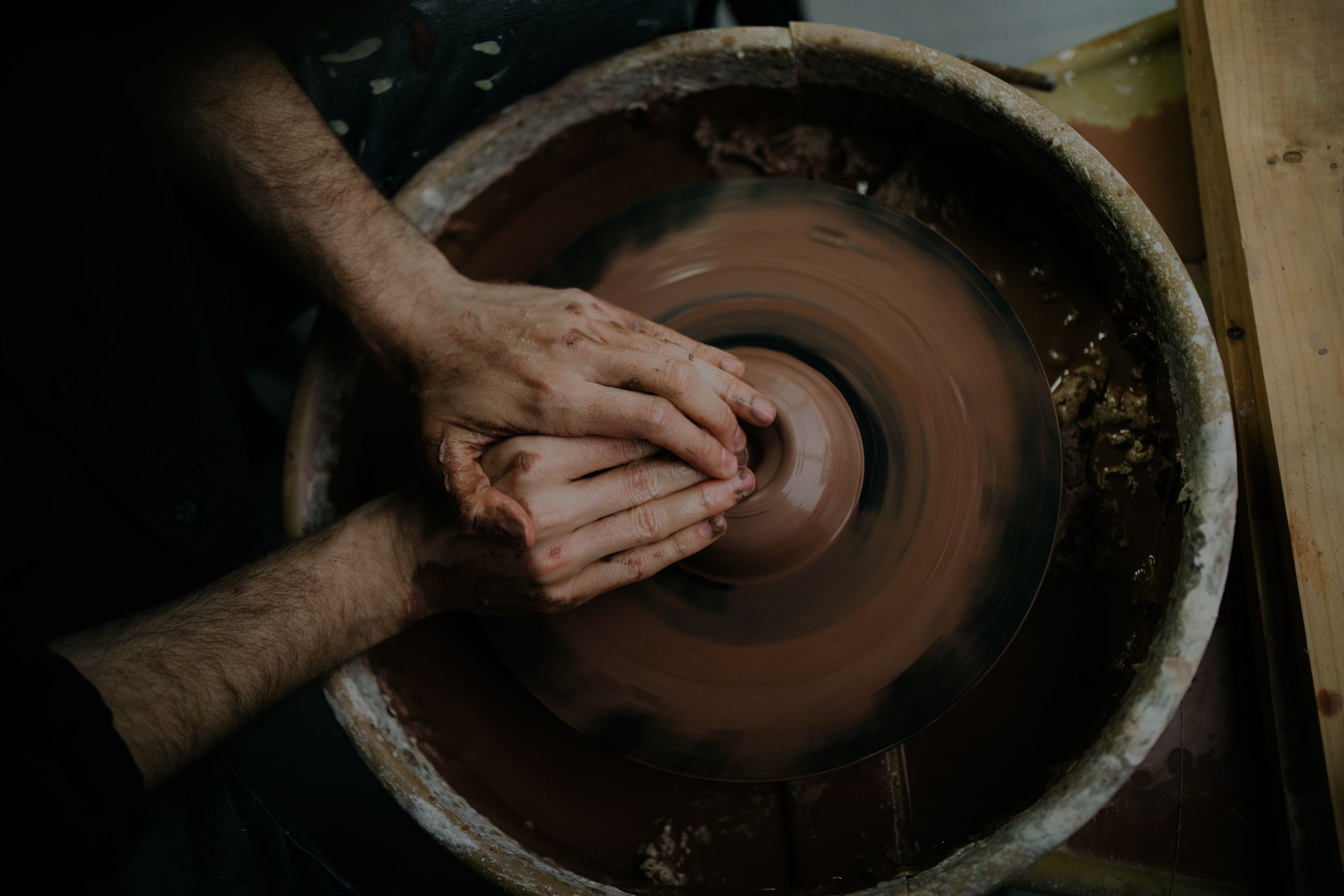A sequence of hands working with clay on a pottery wheel, shaping and forming a vessel from dark brown clay.