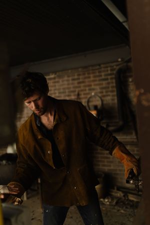 A figure in a brown coat crouches in a dark atmospheric setting against a brick wall background.
