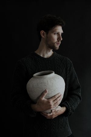 A person wearing a dark sweater holds a large white ceramic vessel against a black background.