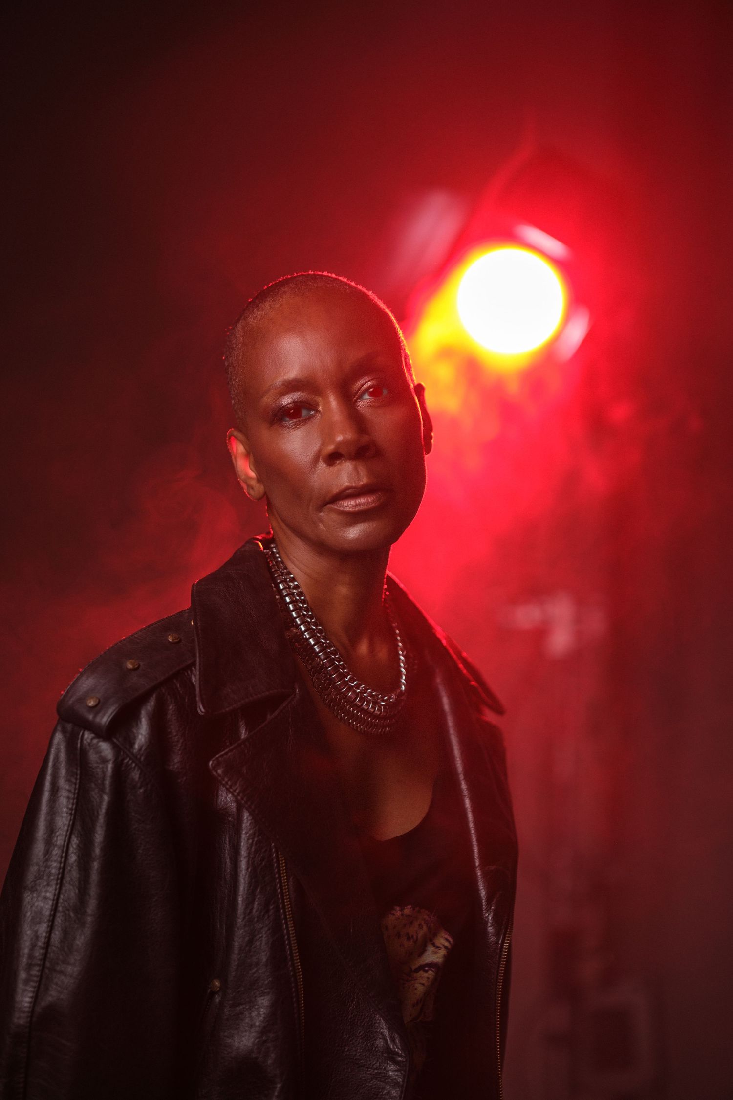 Portrait in dramatic red lighting with leather jacket and necklace against dark background.