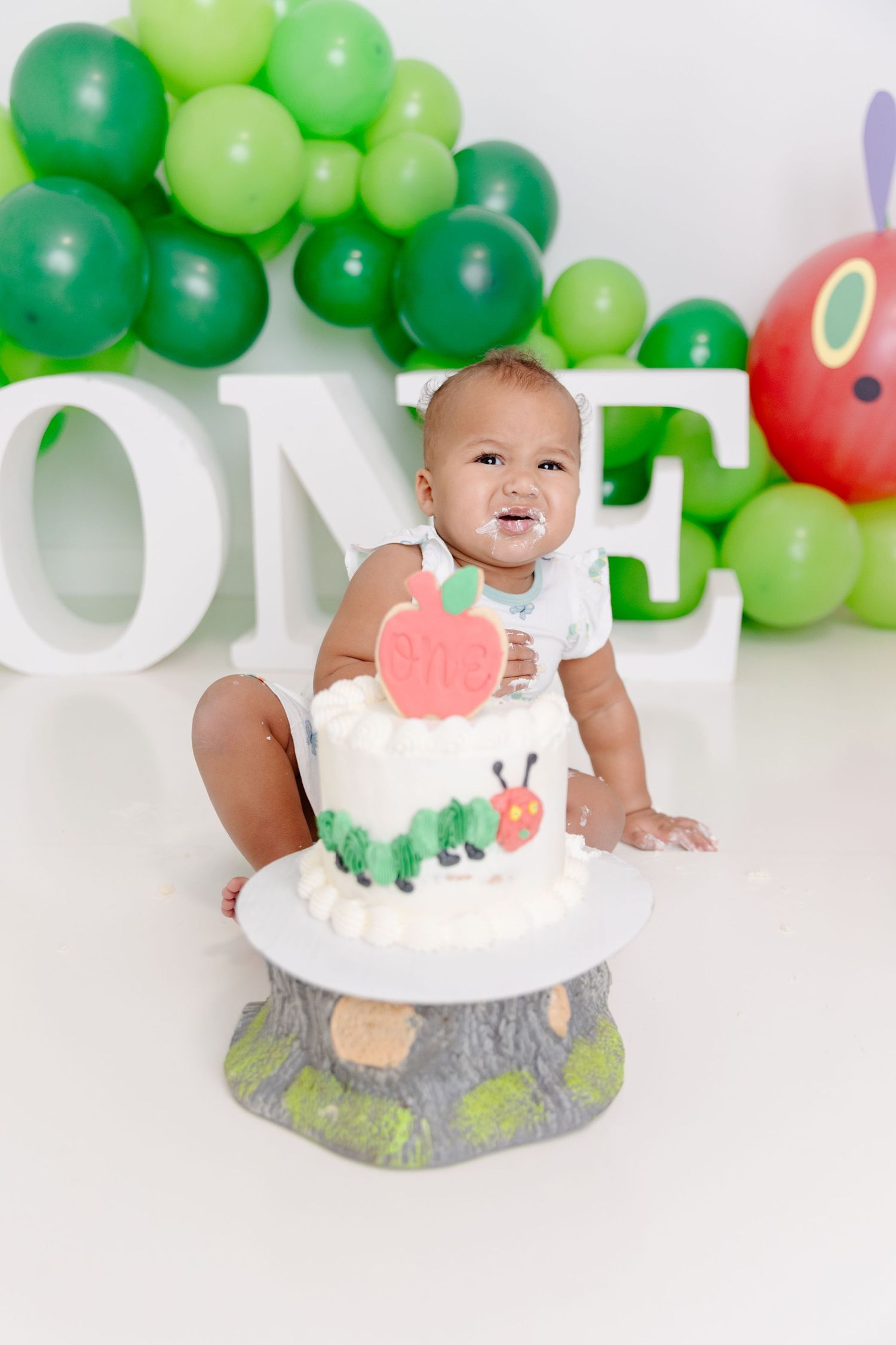 Baby wearing white onesie sits on floor with green balloon decorations and smiles during first birthday cake smash photo session.