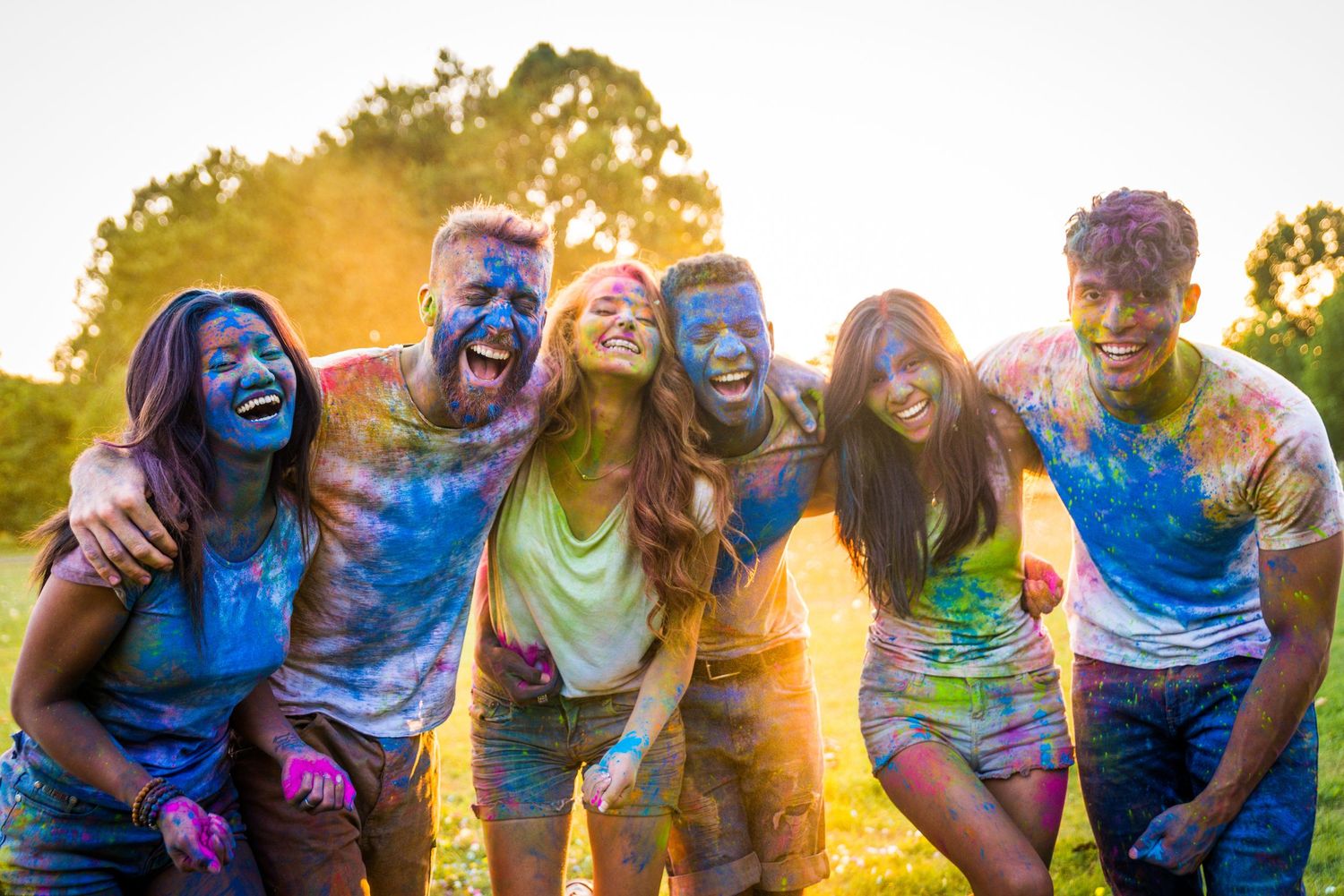 Group of friends covered in colorful powder laugh together at sunset during Holi festival celebration.