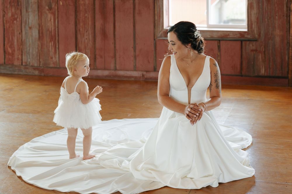 A wedding dress train spreads across rustic wooden floors in a sunlit room as a toddler plays nearby.