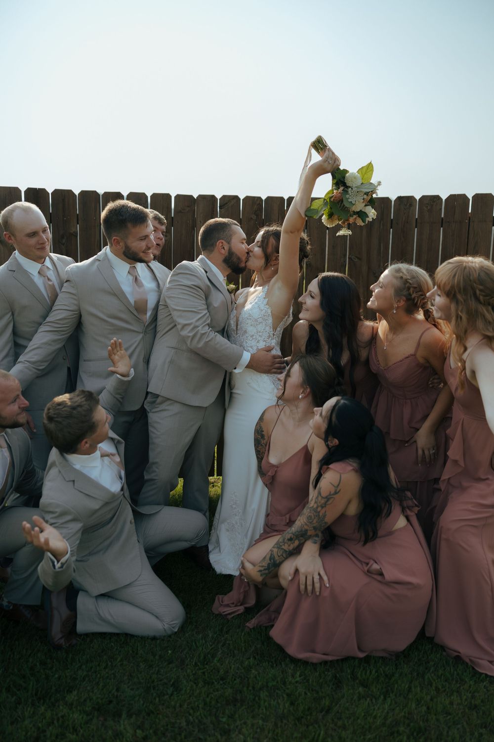 Wedding party in gray suits and mauve dresses pose playfully on grass by wooden fence during outdoor celebration.