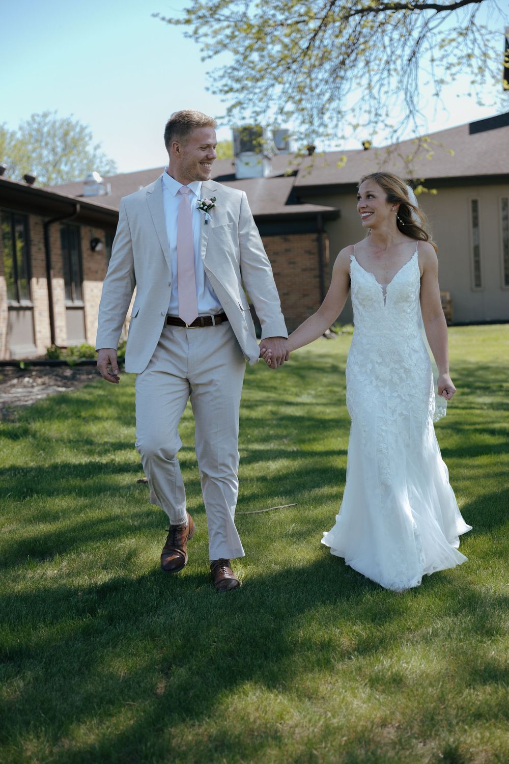 Bride and groom walking together on green grass wearing light colored wedding attire.