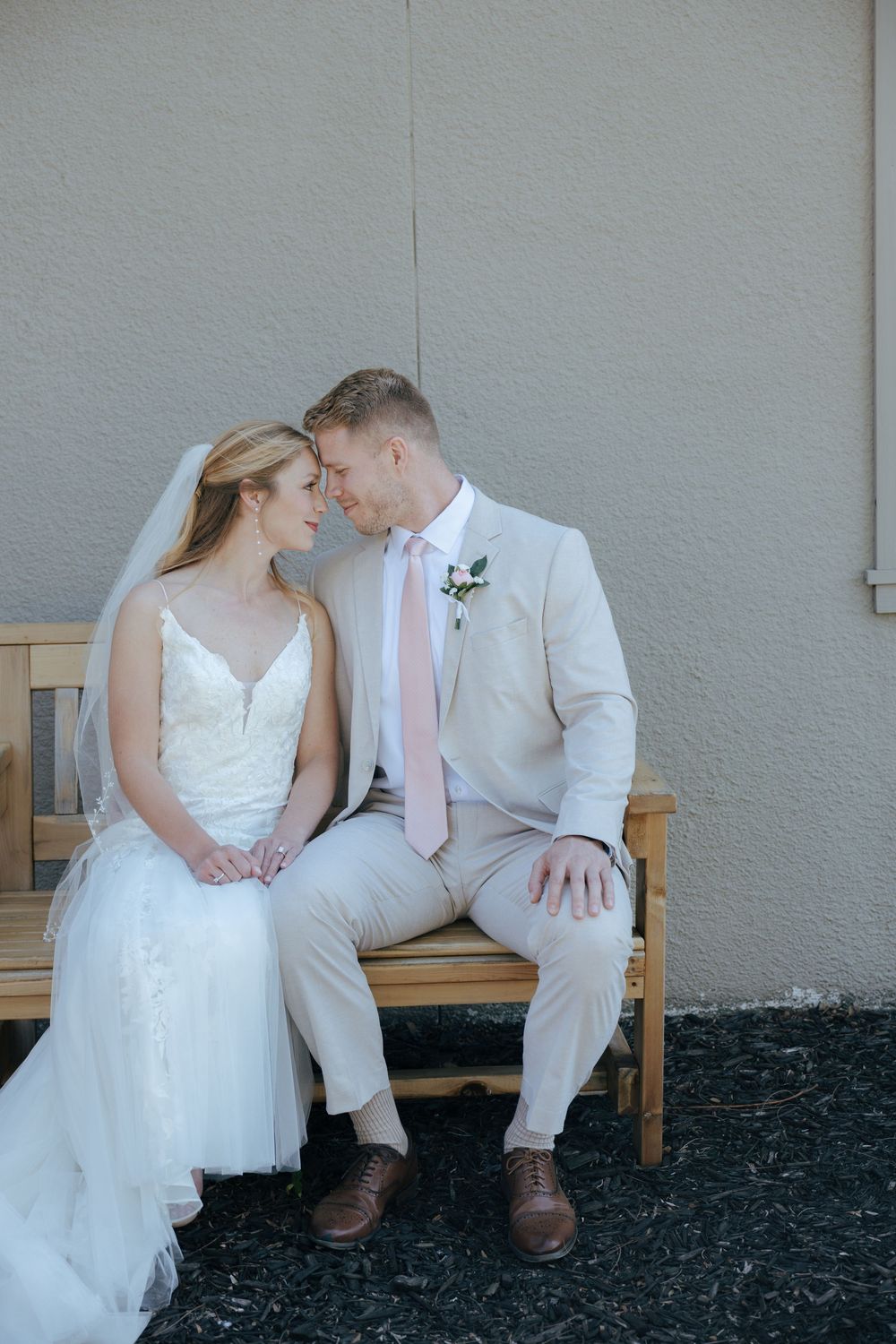 A couple in wedding attire share an intimate moment while sitting on a wooden bench against a gray wall.