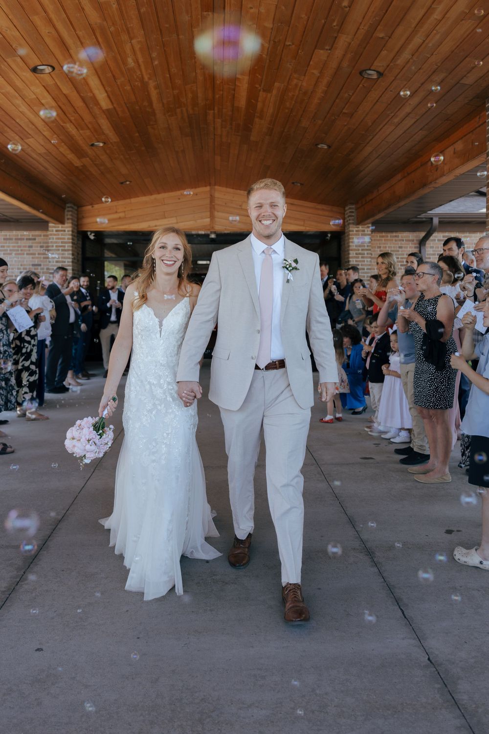 A couple in white wedding attire walks down a covered walkway while guests blow bubbles during their celebration exit.
