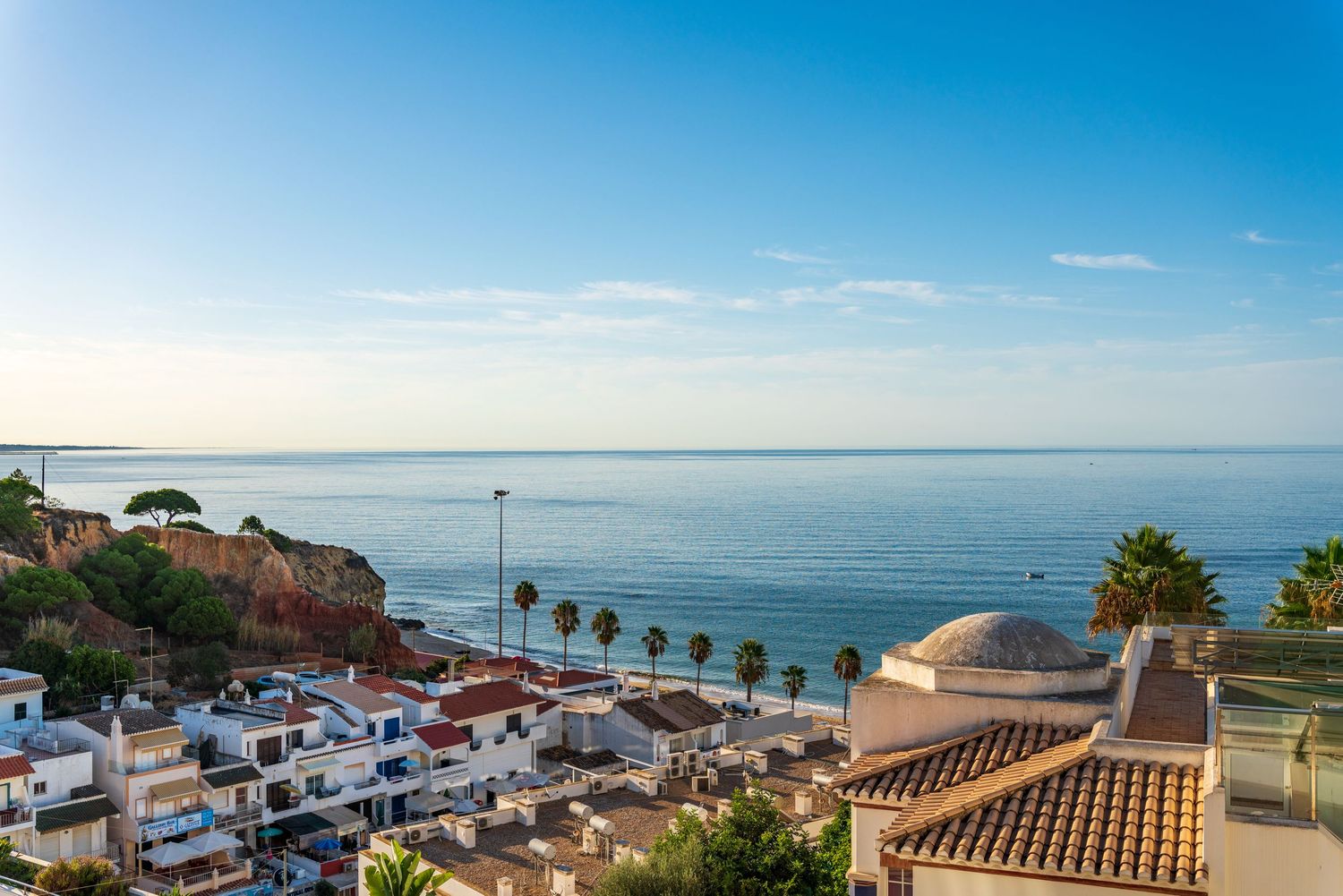 Scenic coastal view of white Mediterranean buildings and palm trees overlooking a calm blue ocean on a sunny day.