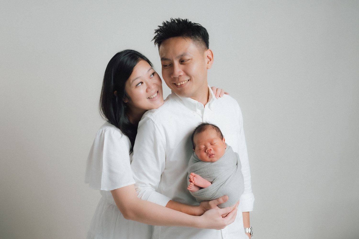 Modern family portrait with newborn baby against light background in matching white outfits.