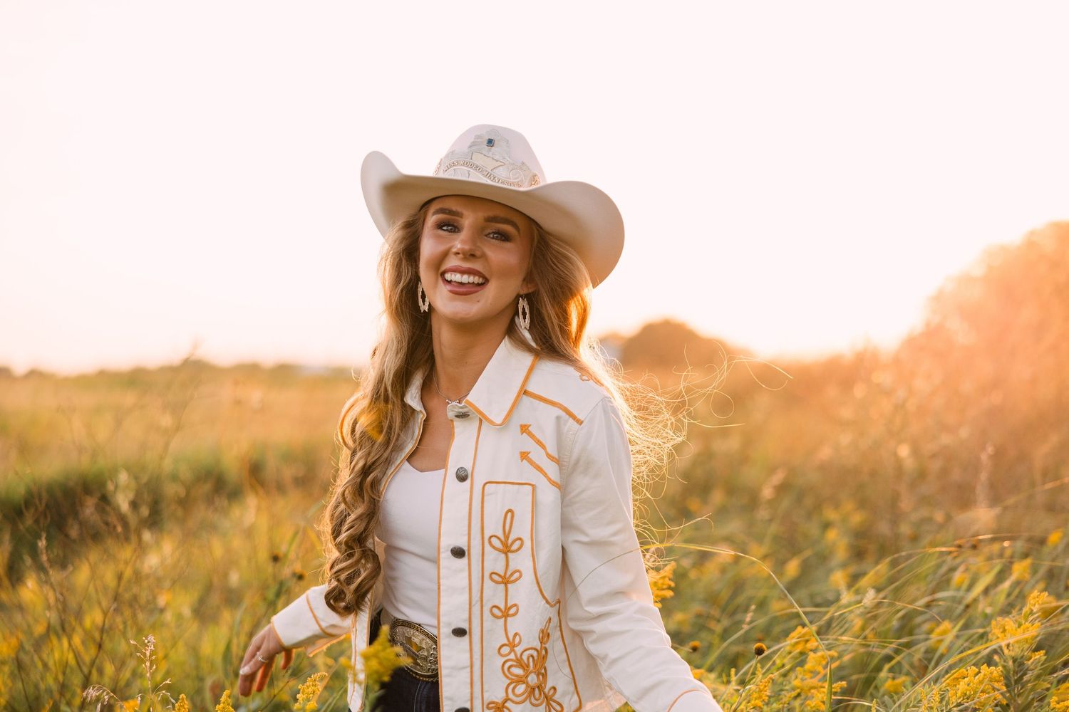 Smiling woman in a white cowboy hat and embroidered western jacket standing in a golden wildflower field at sunset.
