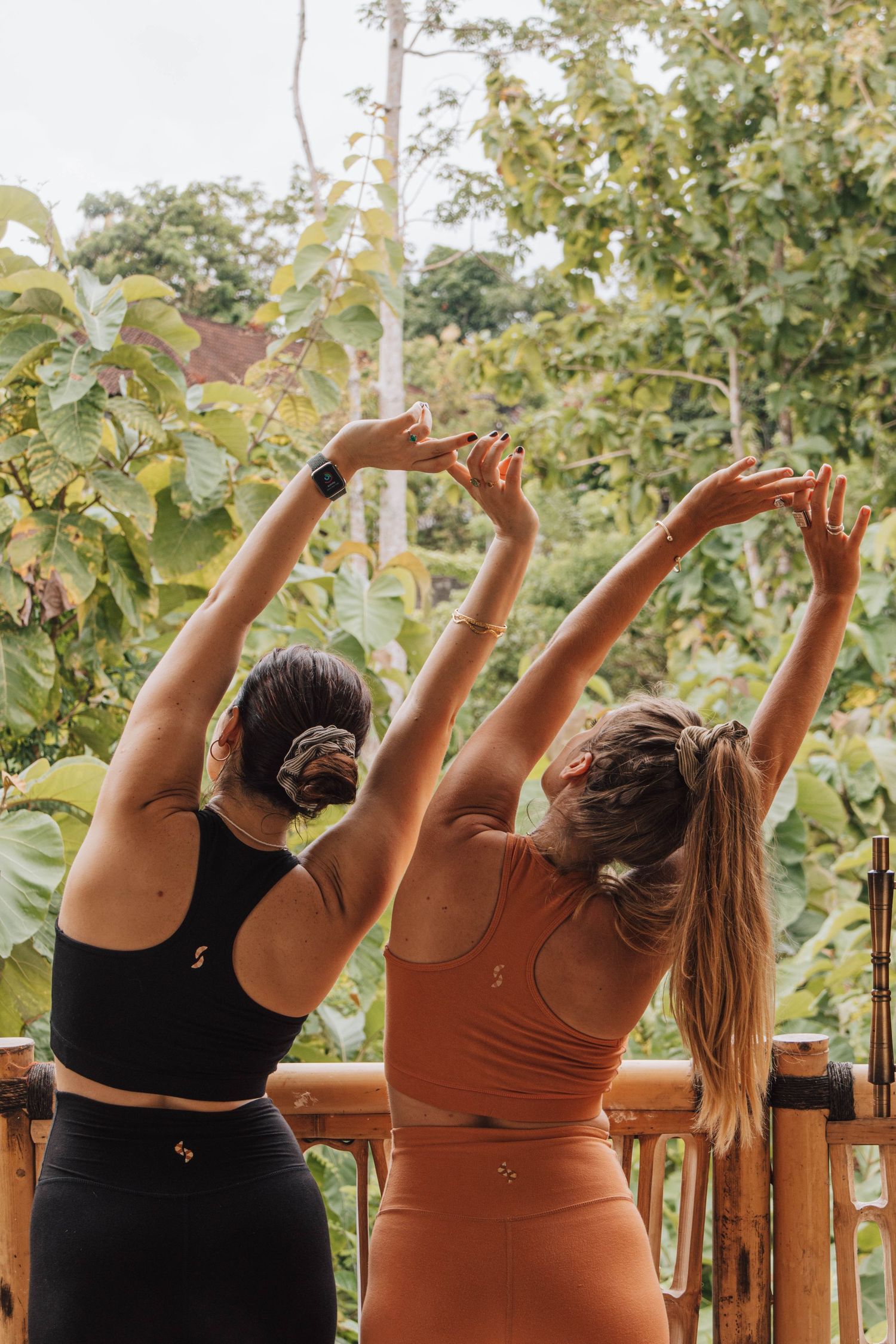 Two women in activewear stretch their arms overhead on a wooden balcony surrounded by lush tropical greenery.