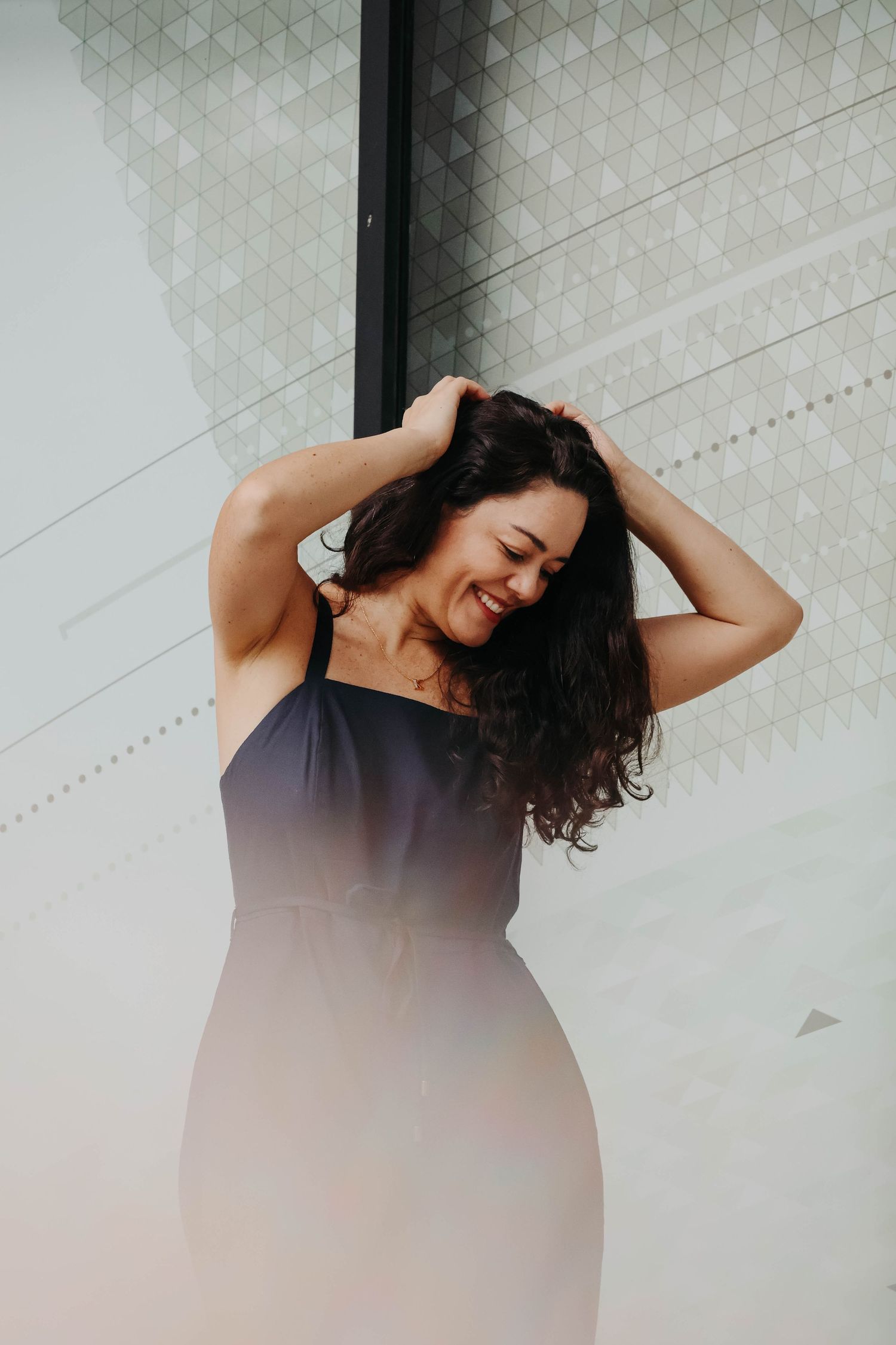 A smiling woman with curly dark hair in a navy dress holds her hair up against a modern geometric wall.