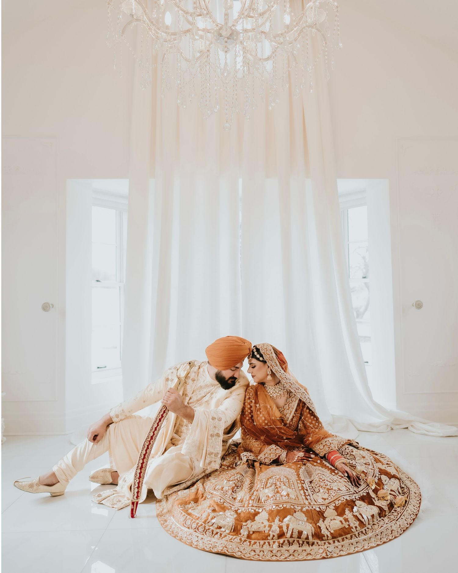 A traditional Indian wedding couple in ornate outfits sit together under an elegant white chandelier.
