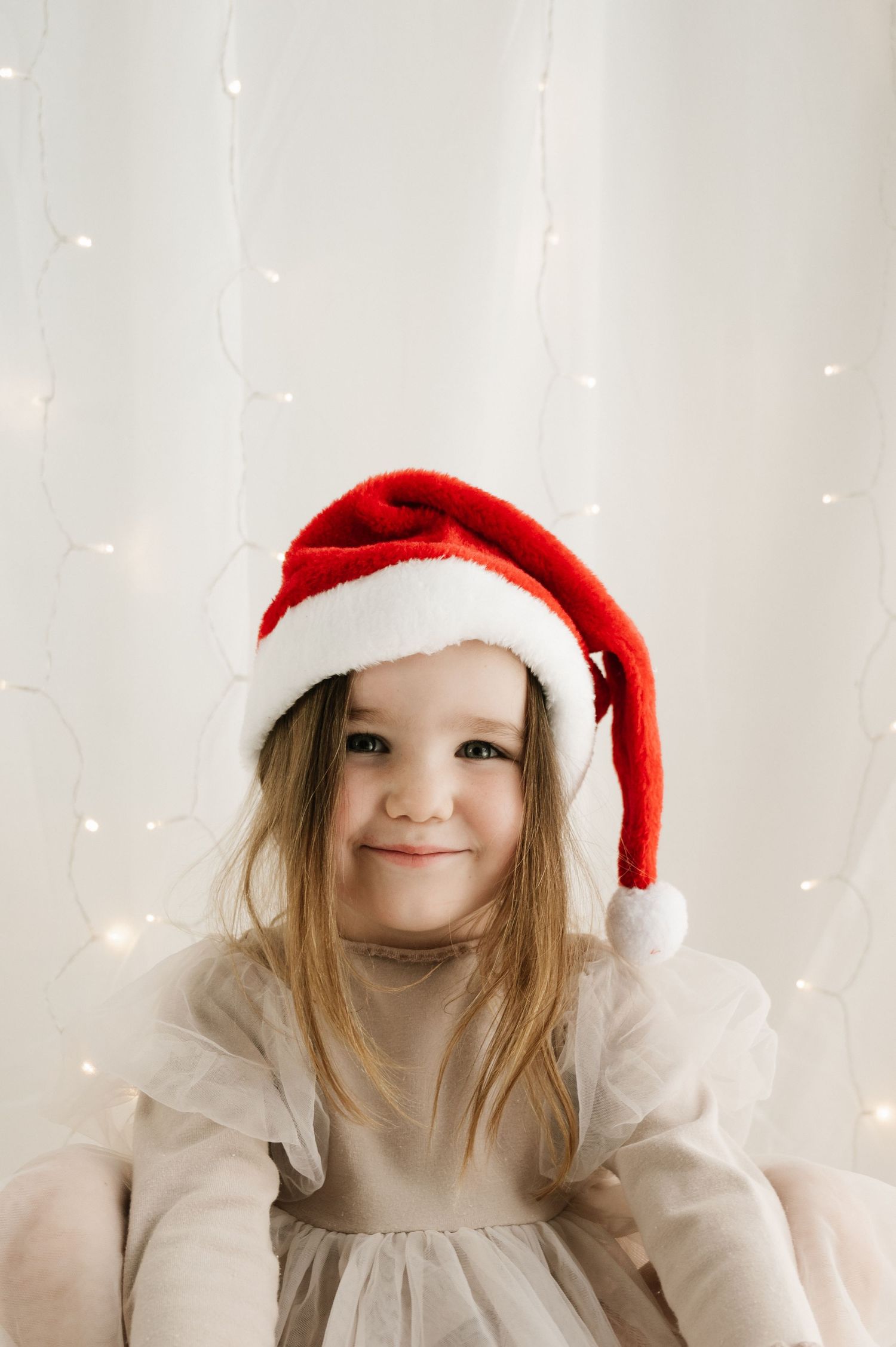 Young child wearing Santa hat and beige dress smiling against white twinkle light background.