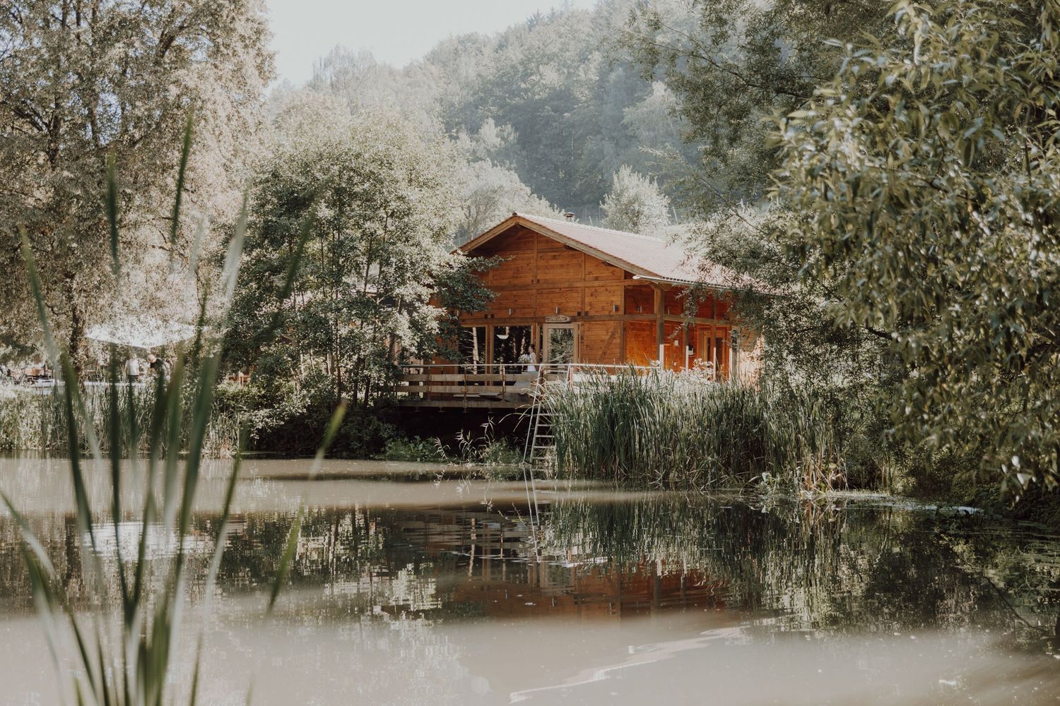 A rustic wooden cabin with a deck reflected in a serene lake surrounded by trees.