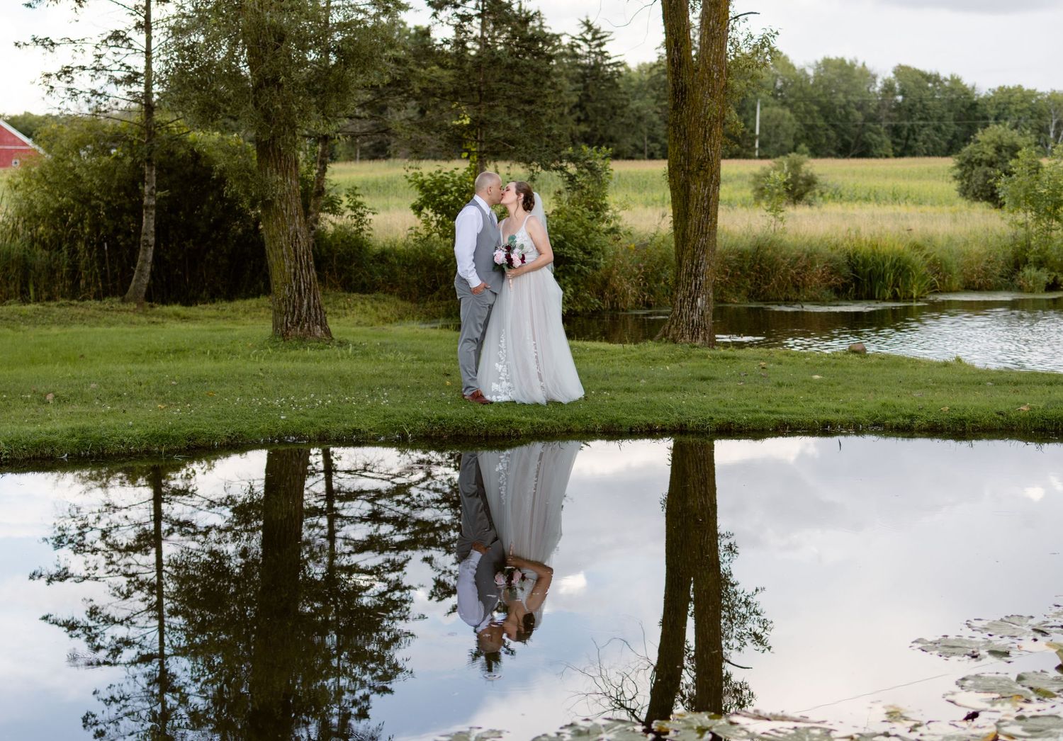 Wedding couple reflected in calm pond water surrounded by trees.