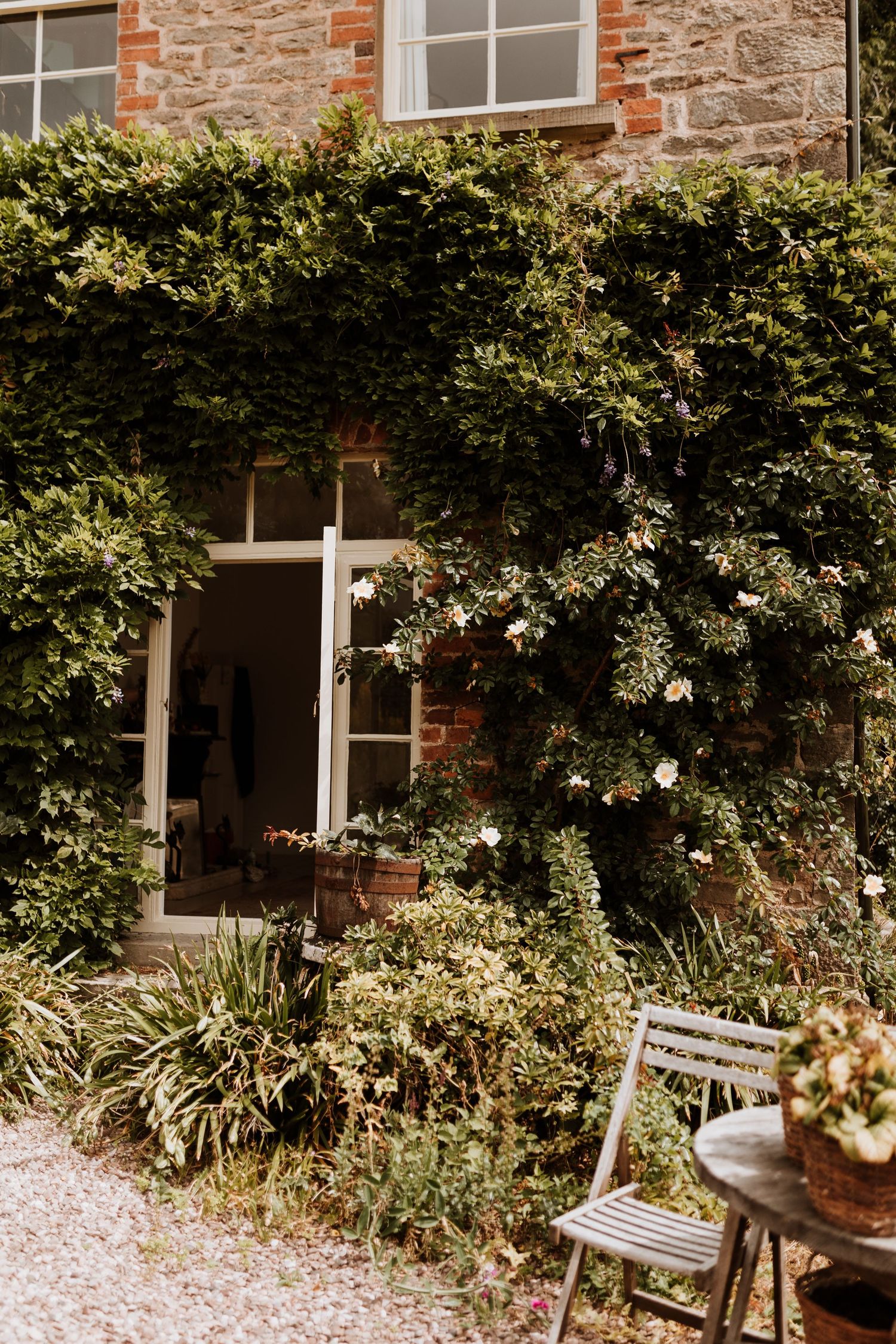 Stone cottage exterior with climbing vines, rustic garden decor and a quaint wooden chair beside a traditional doorway.