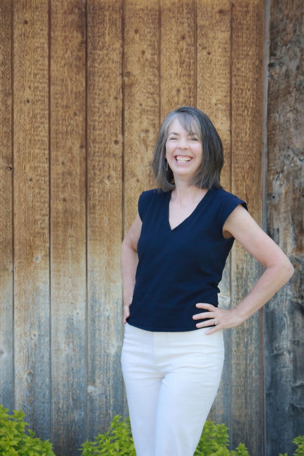 Woman in navy top and white pants poses confidently against wooden wall background.