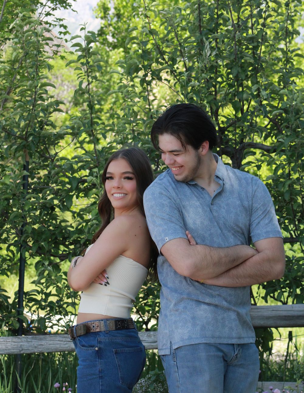 Couple poses casually against lush green foliage with mountains in background.