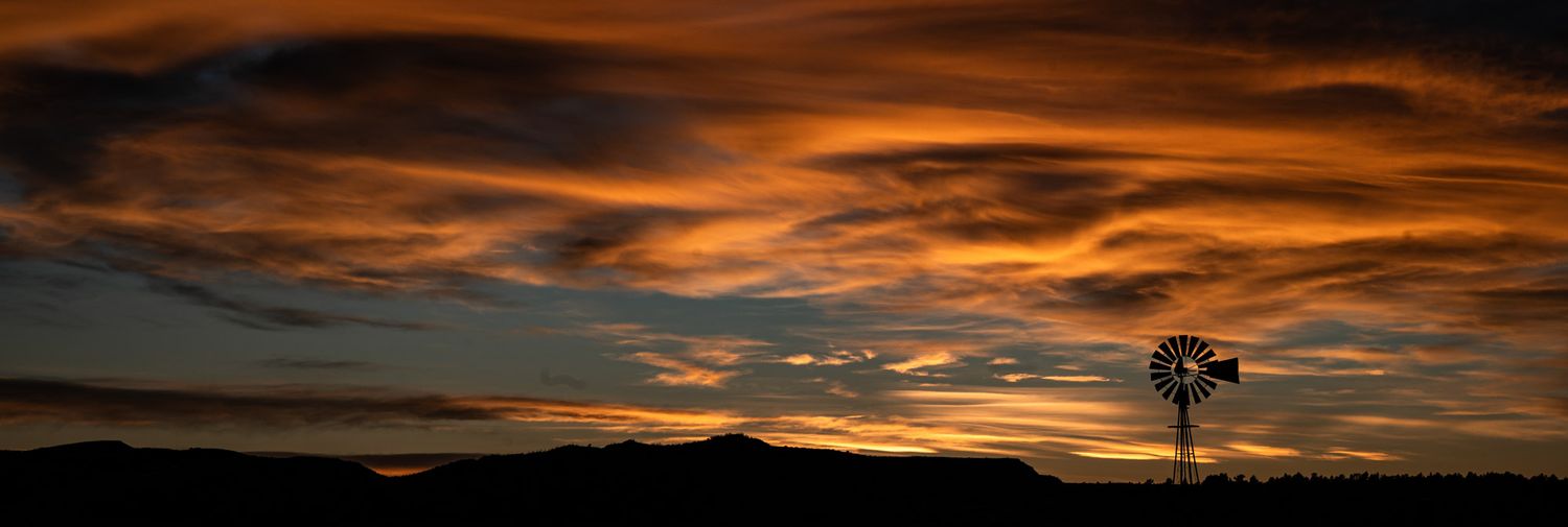 Colorful sunset light illuminates a cloudy sky above silhouetted mountains and a windmill.