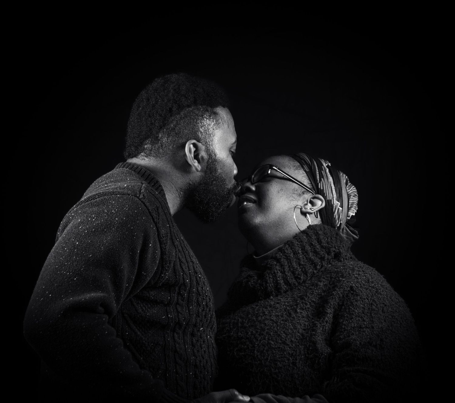 An intimate black and white portrait showing two people in turtleneck sweaters about to kiss against a dark background.