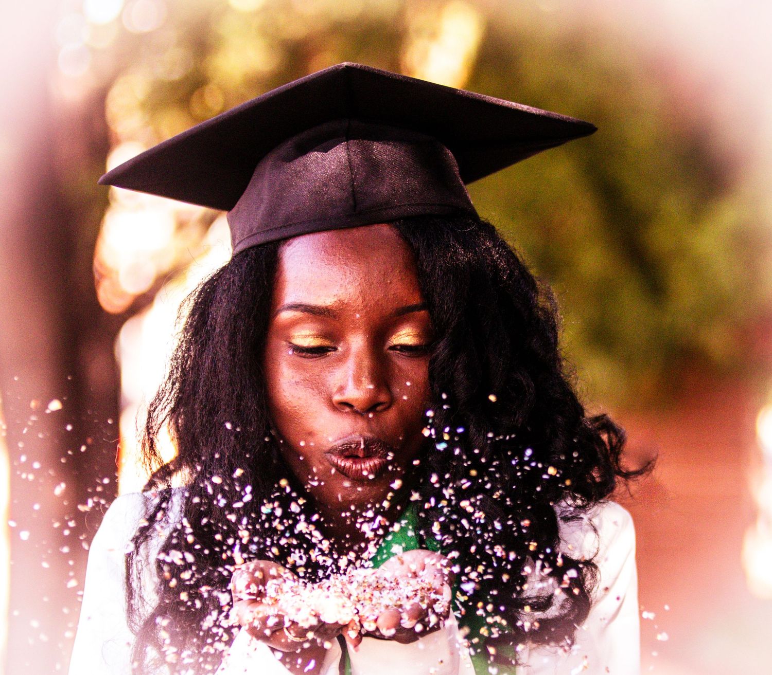 A graduate in cap and gown blows glitter in a dreamy golden-lit portrait.