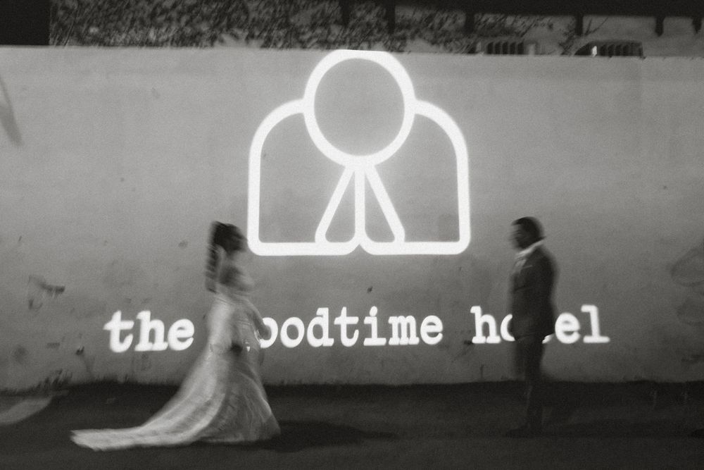 Black and white photo of a bride and groom walking past The Goodtime Hotel in Miami, illuminated sign at night.