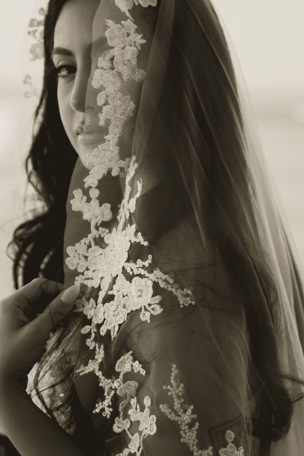 Black and white portrait of bride holding lace-embroidered veil over her face.