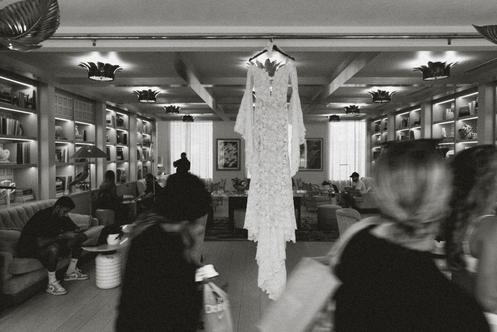 Black and white photo of an elegant lace wedding dress hanging in a stylish library lounge with guests mingling below at The Goodtime Hotel
